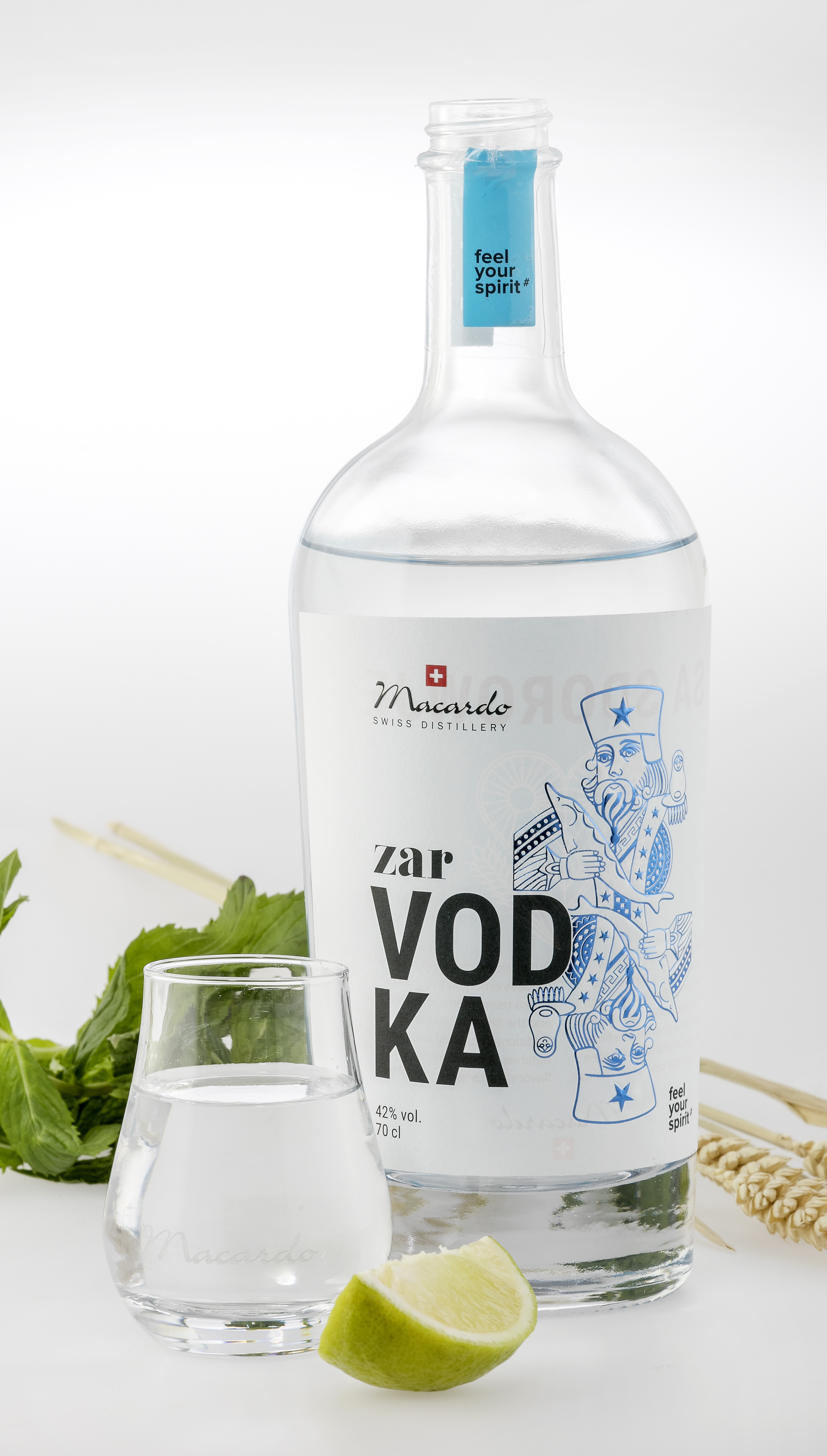 Eine klare Glasflasche Zar Vodka, daneben ein Glas mit dem gleichen Inhalt. Eine Limettenspalte und Minze liegen davor. Im Hintergrund Weizenähren. Das Etikett zeigt den Schriftzug "Macardo Swiss Distillery" und eine blau gezeichnete Zarenfigur.